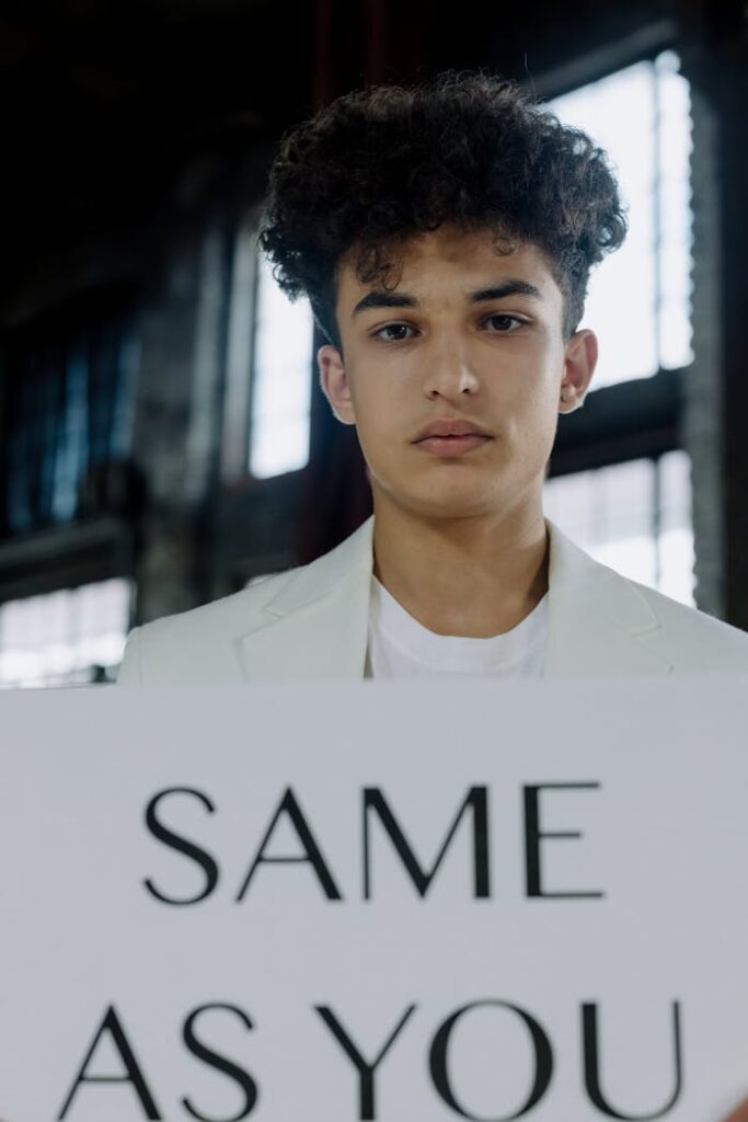 Portrait of a young man showcasing a sign with 'SAME AS YOU' indoors emphasizing equality.